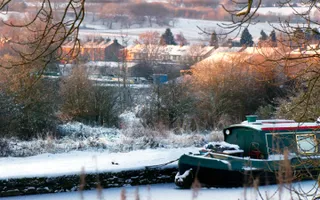 Snow covered countryside and houses surround a narrowboat, snowy towpath, and iced-over canal