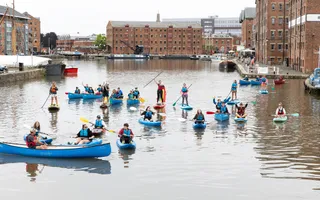 A group of people in multiple blue canoes and paddleboards lift their arms in the air and cheer at the camera, with Gloucester Docks in the background