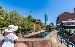 Woman in white clithing looking out onto a canal, standing on a bridge on a sunny day with blue sky and skyscraper in the background