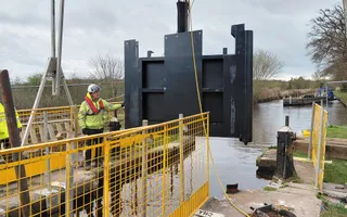 A man in yellow hi vis stands near a black lock gate which is being lowered into a canal.