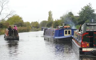 Adults drive narrowboat passed moored boats, with smoke blowing out the chimney