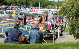 Two men and a dog sit near a line of moored narrowboats at Crick Marina
