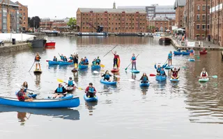 A group of people in blue canoes and paddleboards hold their oars up and smile at the camera with Gloucester Docks buildings in the background