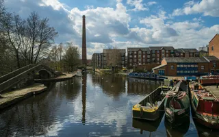 Traditional work boats moor at a three-way canal junction with warehouses to one side and a tall brick chimney in the background.