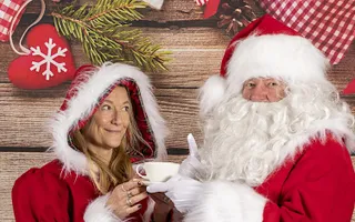 Woman wearing Christmas themed clothing standing next to Santa holding a cup of tea
