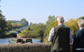 Two adults stand on a bridge looking down at the canal where two narrowboats move through the pound, away from the locks
