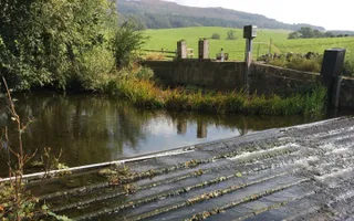 Water runs down a weir at Eshton Beck. A Canal & River Trust worker crouches in the wall in the background