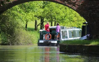 A green and white boat driven by a man in a red t-shirt cruises under a bridge away from camera and round a corner,