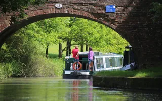 A green narrowboat drives along a scenic canal, passing under a brick bridge, on a sunny day.