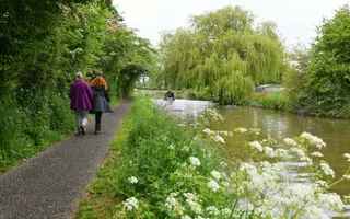 Two people in winter coats walk along a rural towpath, lined with wildflowers, as a narrowboat moves in the distance.
