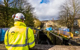 A man in a high vis jacket, helmet, and ear protectors stands on a bridge overlooking an empty lock, where repairs are taking place.