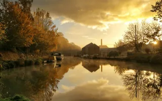 The sun sets over an autumnal canal scene, with boats moored against trees with orange leaves.