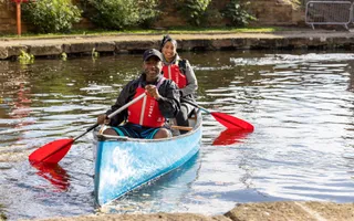 Two people in red life jackets are sitting in a blue canoe holding paddles and smiling at the camera.