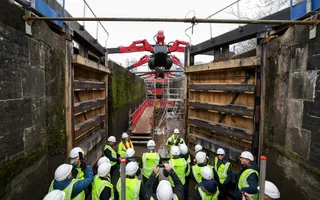 A large number of people in hard hats and hi-vis jackets stand at the bottom of an empty canal to work on a lock gate.
