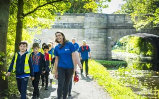 A group of school children with high vis jackets smile as they follow Canal & River Trust employers along the canal towpath on a sunny day.