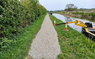 A towpath with fresh gravel is being worked on by a yellow and black machine attached to a boat in the water.