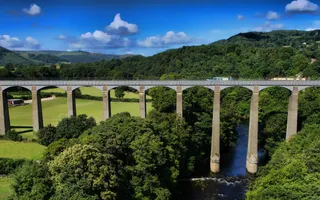 Two narrowboats moving along a canal aqueduct over a river with fields and trees below, and hills and blue skies behind