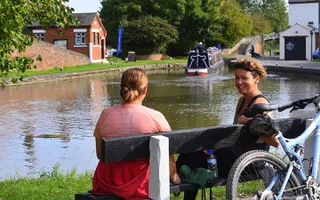 Two women on a bench with a bike in front of the canal, where a boat moves into a lock behind.