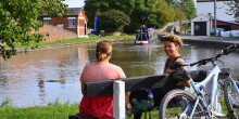 Two seated young women chat next to the canal with a blue bike nearby
