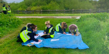 A group of young people studying outside sat on a tarpaulin on the grass next to a canal